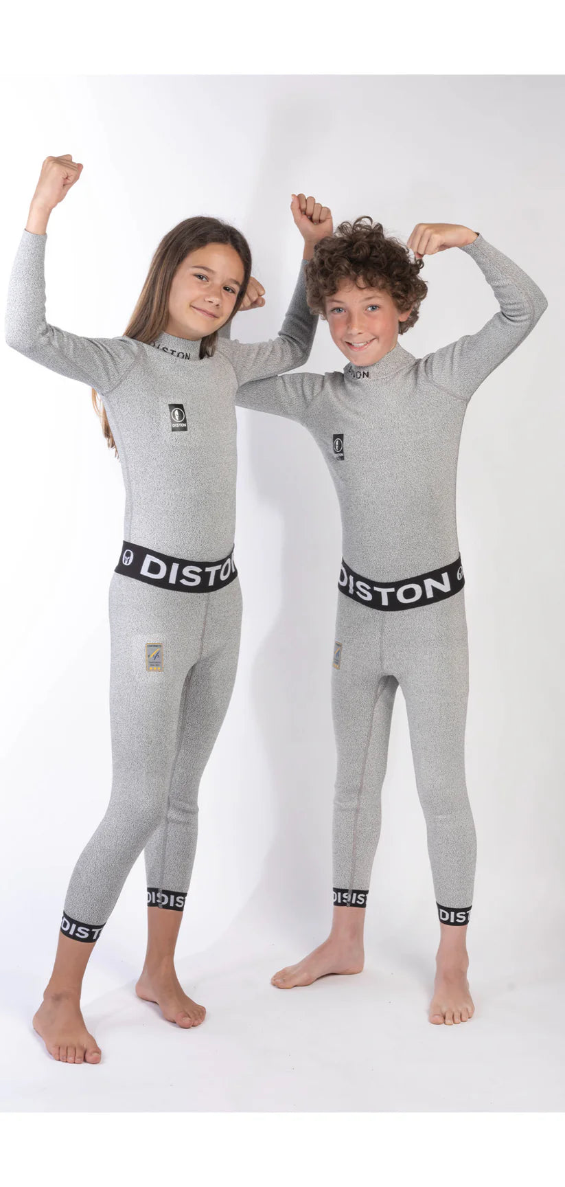 Zwei Kinder in passenden Diston DISTON Cut Resistant Unterhosen Junior - 4FIS Stars in CRG - CORE / Racing Fit stehen lächelnd und federnd nebeneinander - ideal als schnittfeste Unterwäsche oder Schutzunterwäsche Ski für selbstbewusste, aktive Tage.