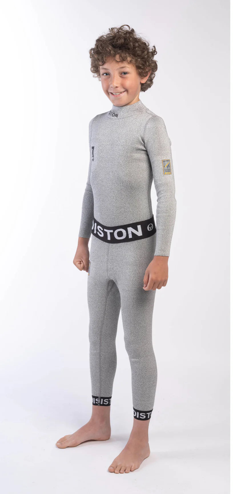 Ein lächelnder, lockiger Junge steht barfuß in einer hellgrauen Diston Cut Resistant Underpants Junior-4FIS Stars in CRG, CORE/Racing Fit mit schwarzen "DISTON"-Bändern an der Taille und an den Knöcheln vor einem schlichten weißen Hintergrund.