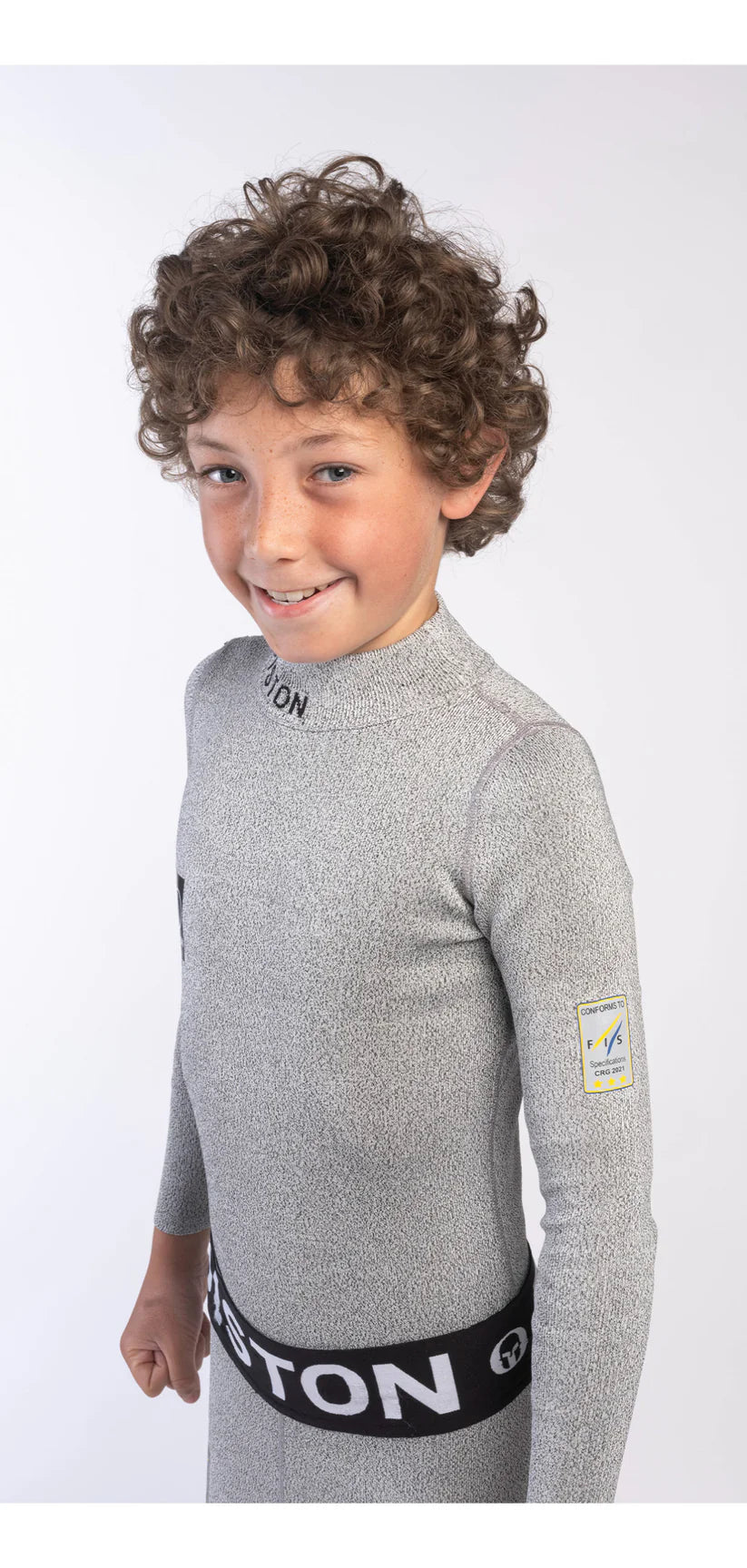 Ein lächelnder Junge mit lockigem Haar modelliert die Diston DISTON Cut Resistant Unterhose Junior - 4FIS Stars in CRG - CORE / Racing Fit, gepaart mit einem langärmeligen grauen Oberteil mit Logoaufnäher und schwarzem Band auf weißem Grund.