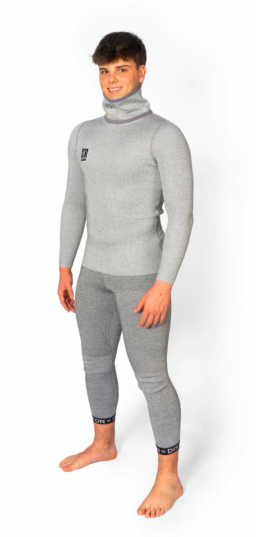 Ein junger Mann steht barfuß, lächelnd, in einem taillierten Diston DISTON WC Cut Resistant Top Men - 4FIS Stars in CRG - CORE / Racing Fit. Das hellgraue Outfit wirkt weich und warm - perfekt für Skirennläufer. Der Hintergrund ist einfarbig weiß.