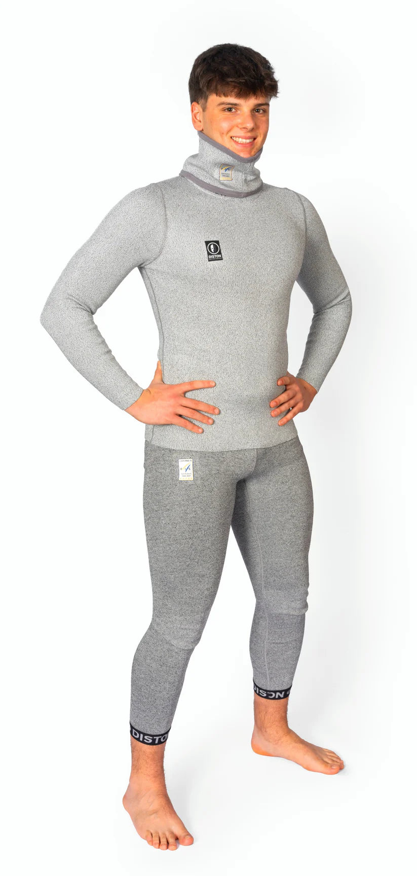 Eine lächelnde Person steht barfuß mit den Händen in den Hüften und trägt ein passendes graues Diston DISTON WC Cut Resistant Top Men - 4FIS Stars in CRG - CORE / Racing Fit und eine für Wasseraktivitäten konzipierte Hose vor einem weißen Hintergrund.
