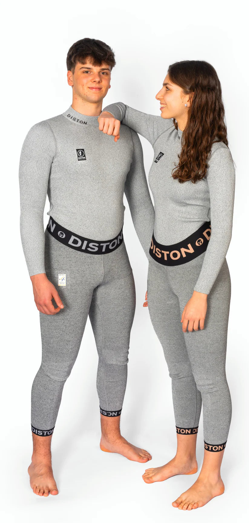 Zwei Personen stehen barfuß in passender hellgrauer Diston-Sportbekleidung. Die Frau, die lächelnd den Arm auf die Schulter des Mannes legt, trägt das DISTON WC Cut Resistant Top Women - 4FIS Stars in CRG - CORE / Racing Fit, inspiriert von den Basisschichten des Skirennsports.