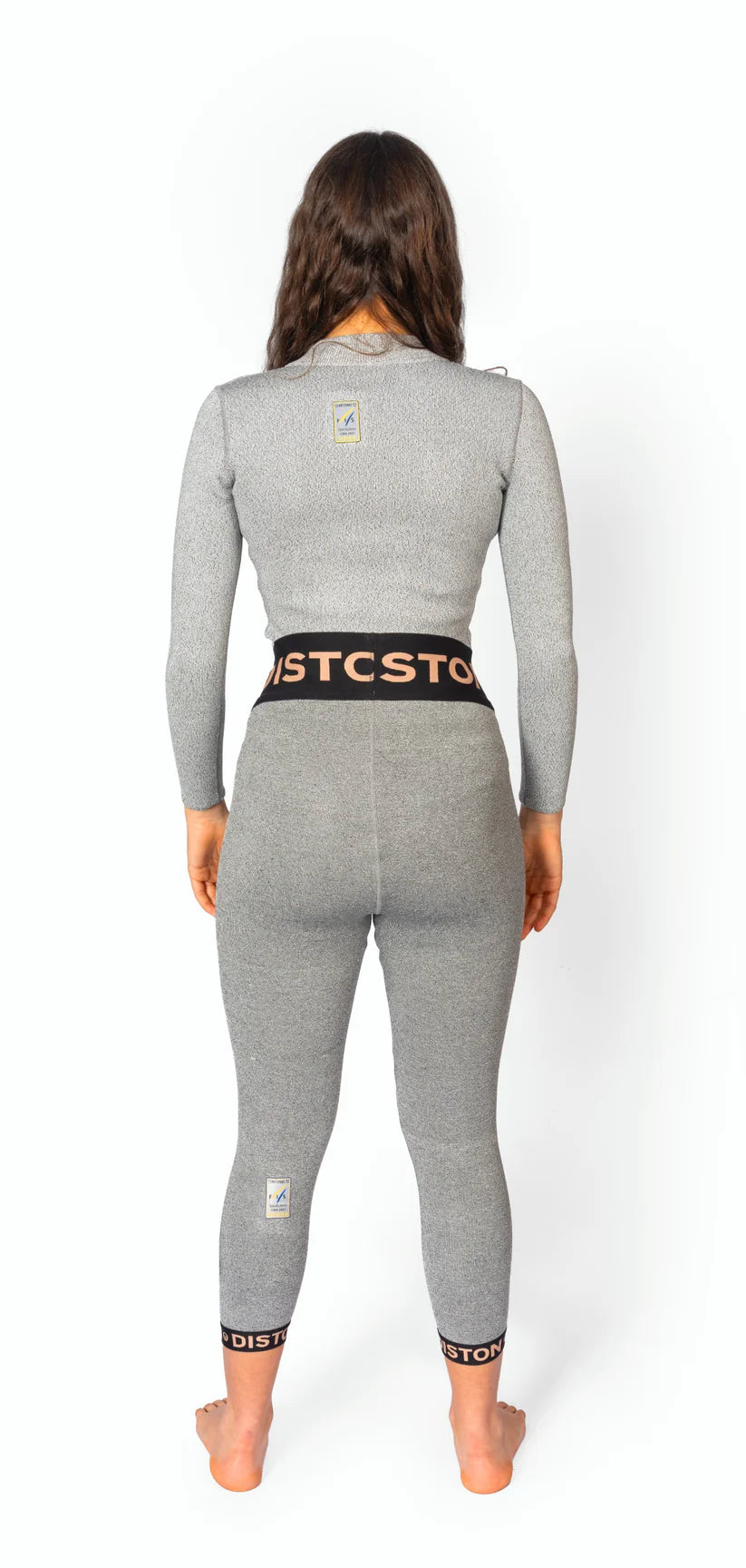Eine Frau mit langen braunen Haaren steht barfuß, den Blick abgewandt, und trägt eine graue Diston DISTON WC Cut Resistant Unterhose für Frauen mit schwarzen und orangefarbenen Streifen und kleinen Logoaufnähern auf dem Rücken.