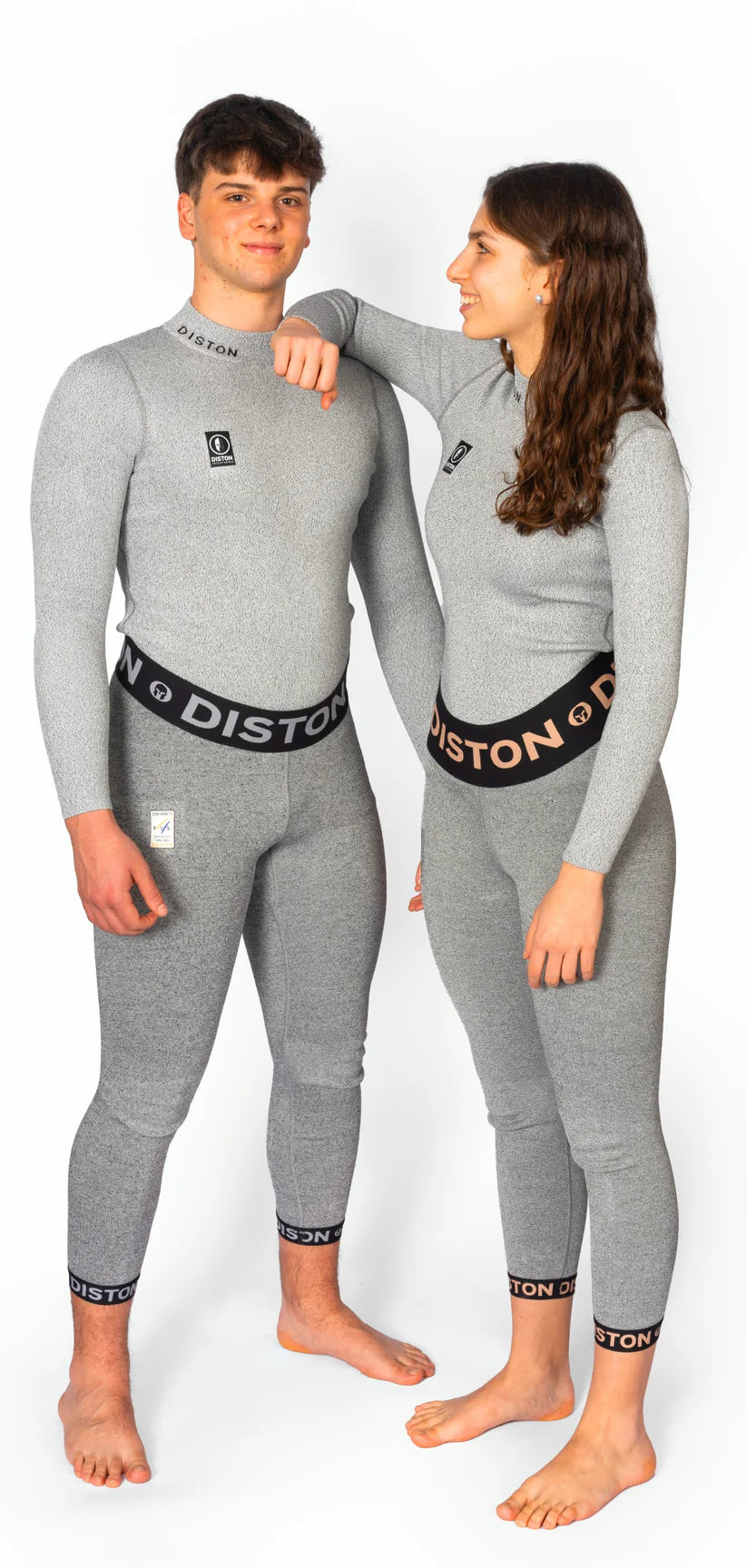 Zwei Personen stehen barfuß vor einem weißen Hintergrund, beide in passenden grauen Diston Racing Compression Fit Anzügen. Die Frau trägt lächelnd eine DISTON WC Cut Resistant Unterhose (Women - 5FIS Stars in CRG - PRO) und stützt ihren Arm auf die Schulter des Mannes.