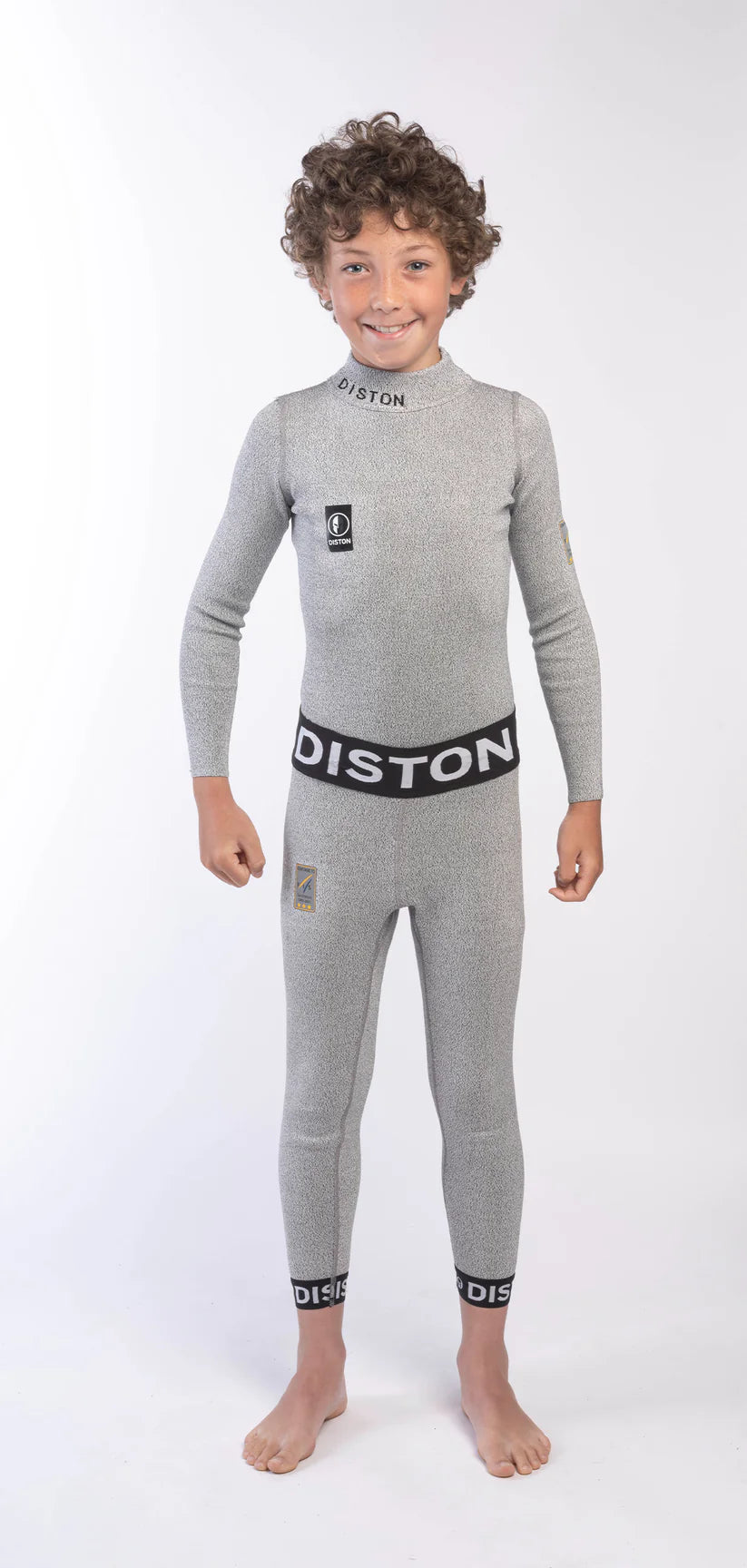 Ein lächelndes Kind steht barfuß auf weißem Untergrund und trägt die DISTON Cut Resistant Unterhose Junior - 3FIS Stars in CRG - BASE / Performance Fit-ideal für junge Skirennfahrer, die schützende und bequeme Funktionskleidung suchen.