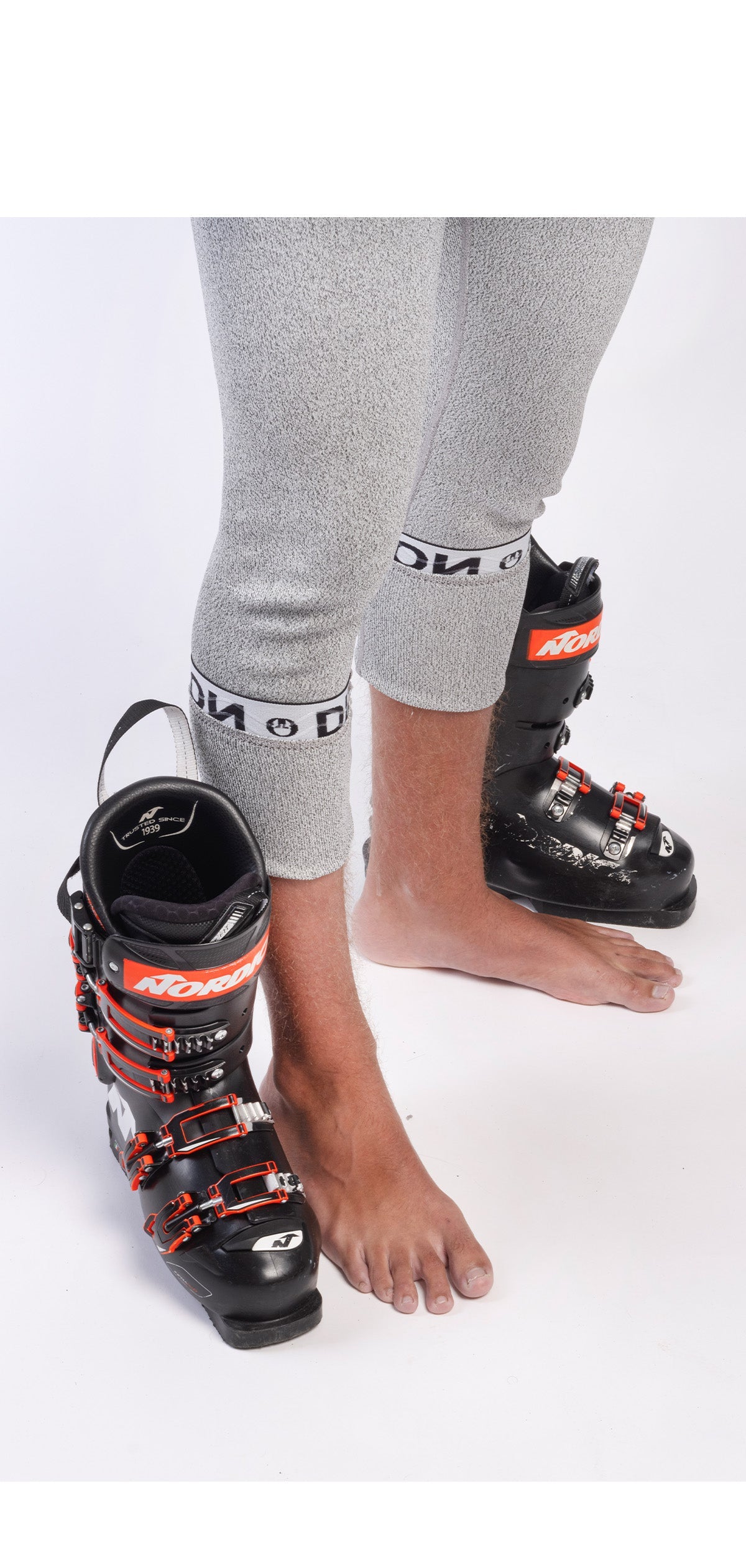 Eine Person, die Diston DISTON WC Schnittfeste Hosen Herren - 4FIS Stars in CRG - CORE/Racing Fit graue Leggings trägt, steht barfuß, mit einem Fuß neben einem schwarz-roten Skischuh und mit dem anderen in einem identischen Schuh.