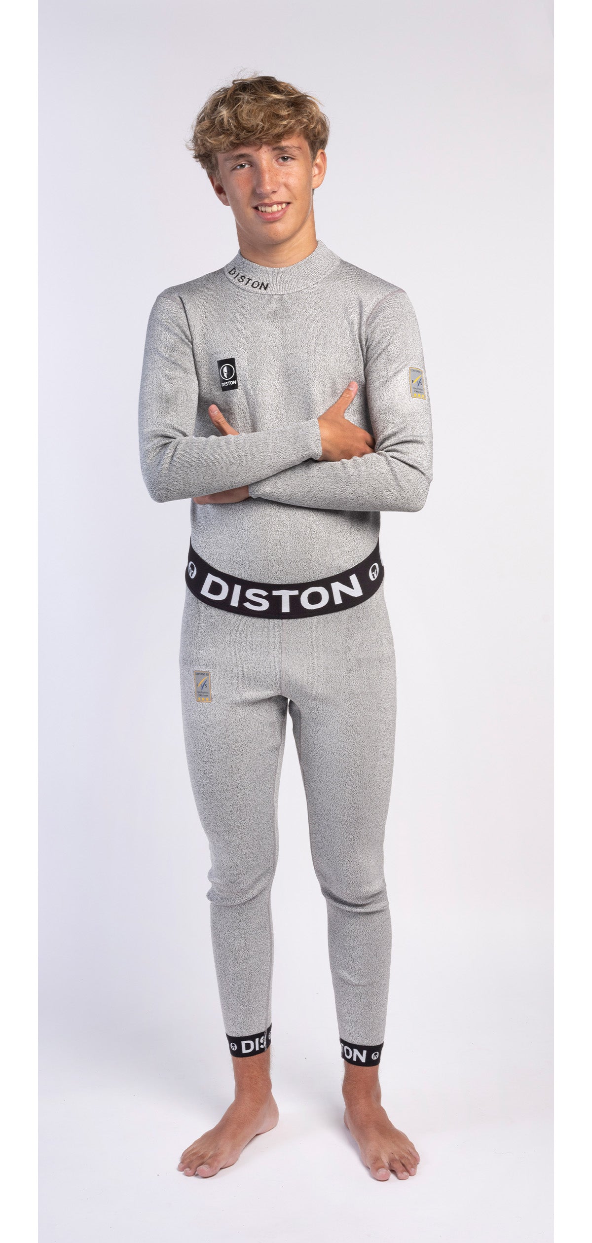 Ein junger Mensch mit kurzen, lockigen Haaren steht barfuß und lächelnd vor einem weißen Hintergrund und trägt die DISTON WC Schnittfeste Hosen Herren - 4FIS Stars in CRG - CORE / Racing Fit von Diston als schützende Schnittschutzunterhose.