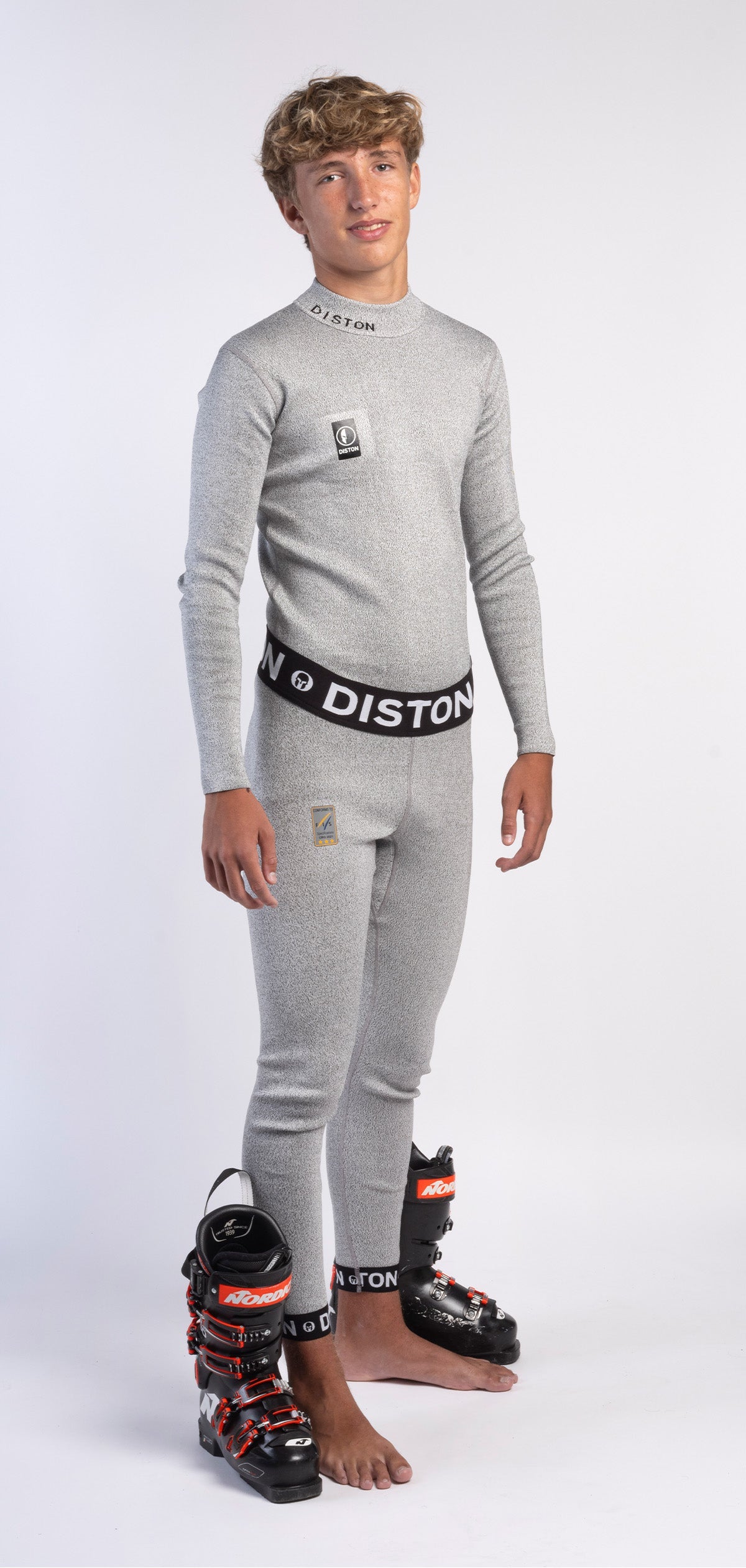 Eine junge Person mit kurzen, hellbraunen Haaren trägt einen Diston WC Schnittfeste Hosen Herren - 4FIS Stars in CRG - CORE / Racing Fit Base Layer, barfuß, mit schwarzen und roten Skischuhen vor sich.