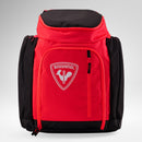 Eine rot-schwarze Rossignol HERO ATHLETES BAG mit einer Fronttasche mit Reißverschluss und dem ikonischen Rossignol-Logo, das einen Hahn auf einem Schild zeigt. Sie eignet sich ideal als Rucksack für die Skirennausrüstung und steht aufrecht vor einem hellgrauen Hintergrund.