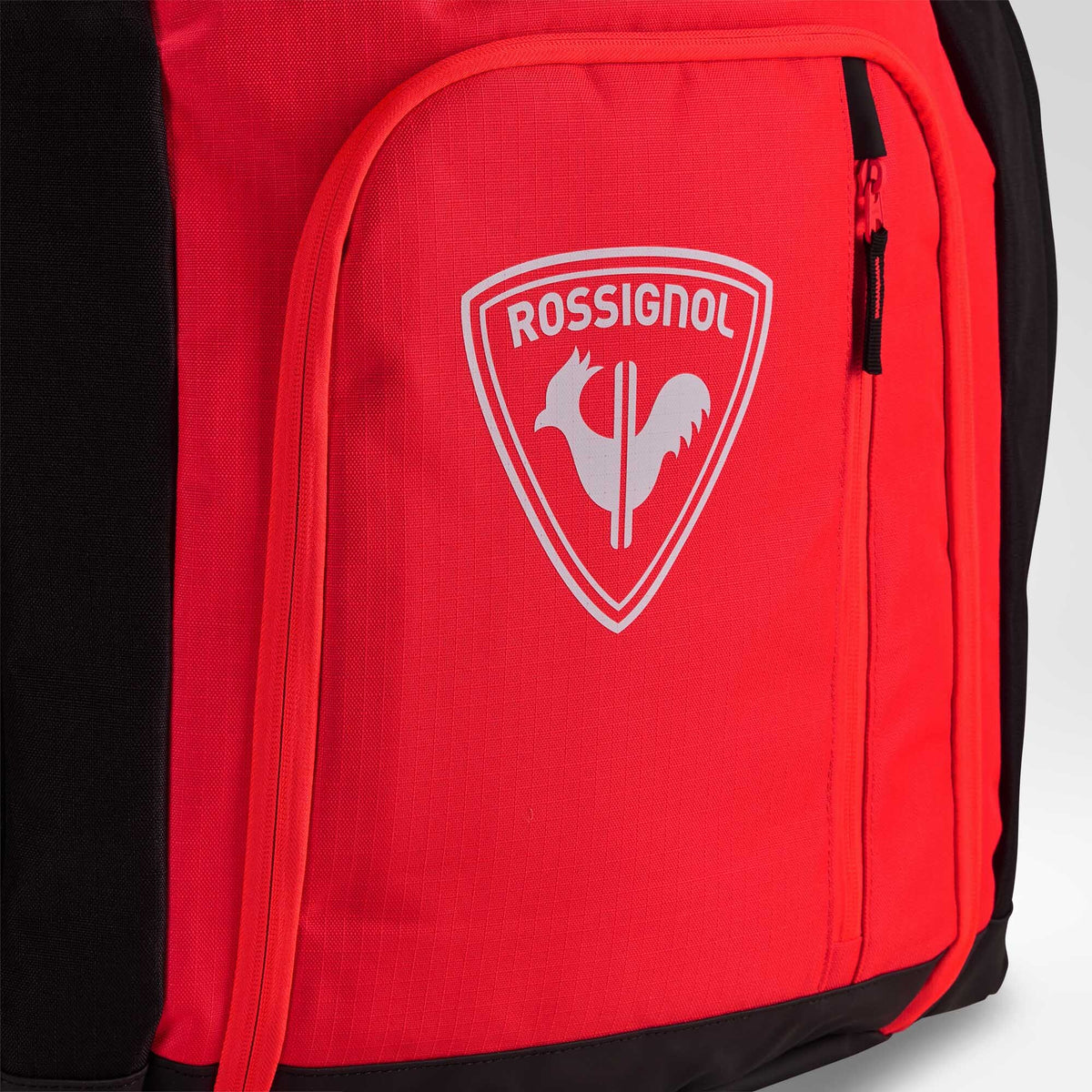 Nahaufnahme einer roten Rossignol HERO ATHLETES BAG mit einer Reißverschlusstasche, auf deren Vorderseite das weiße Rossignol-Logo und das Hahn-Emblem zu sehen sind.
