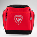 Eine leuchtend rote Rossignol HERO HEATED BAG 230V mit dem ikonischen weißen Hahn-Logo, einem robusten schwarzen Boden, praktischen Seitentaschen sowie zwei Reißverschlusslaschen mit den Aufschriften „230V“ und „12V“, sticht vor einem hellen Hintergrund mit Farbverlauf hervor – ideal für den Ski-Weltcup oder zum Warmhalten Ihrer Skiausrüstung dank integrierter Schuhheizung.