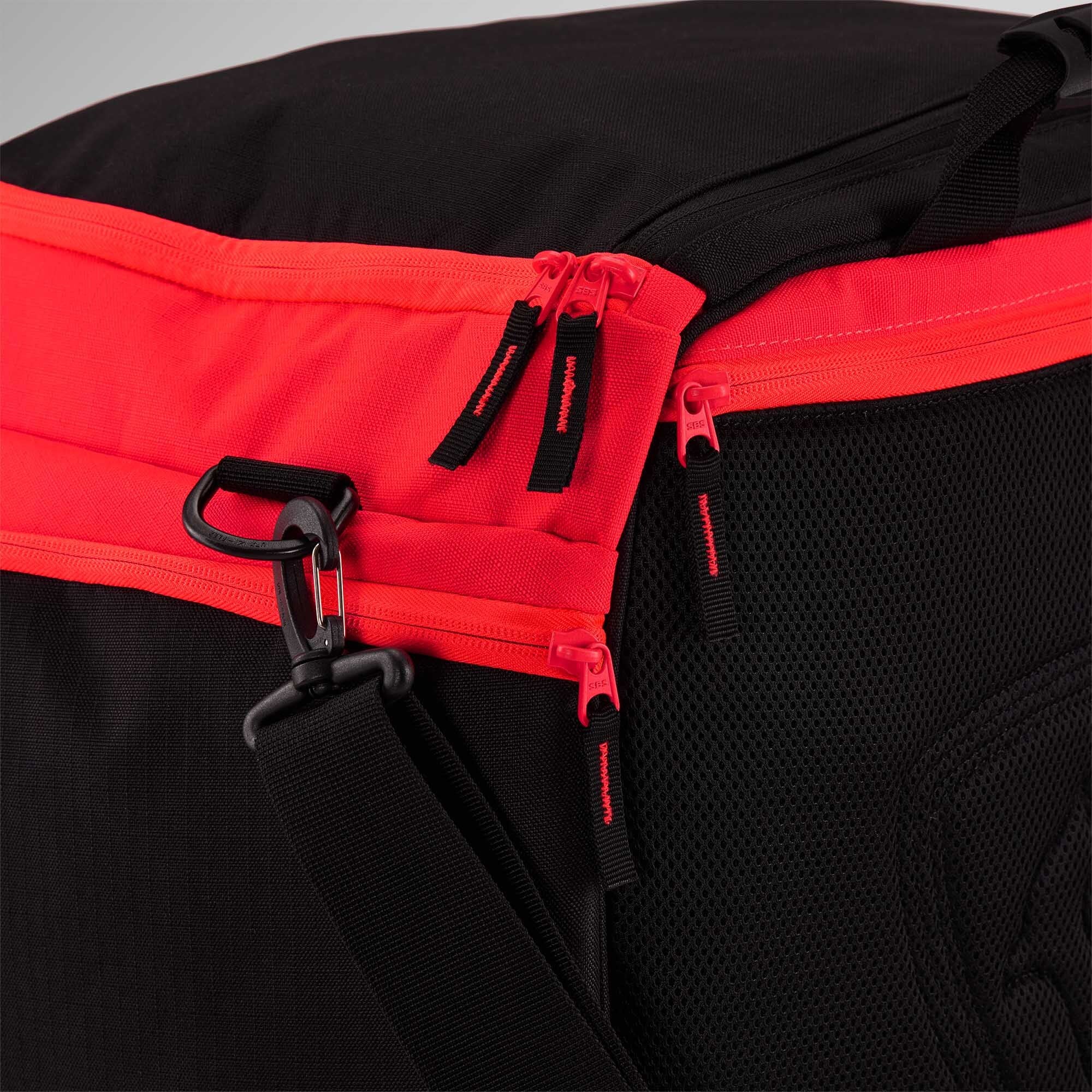 Nahaufnahme einer schwarzen Rossignol HERO HEATED BAG 230V mit einem leuchtend roten Reißverschluss, drei Reißverschlusszügen und einem schwarzen, verstellbaren Schultergurt, der an einem Metallring befestigt ist. Dank ihrer Konstruktion aus Netzgewebe und Stoff eignet sich die Tasche ideal, um Ihre Skischuhe warm zu halten – dank des integrierten Skischuh-Heizsystems von Rossignol.