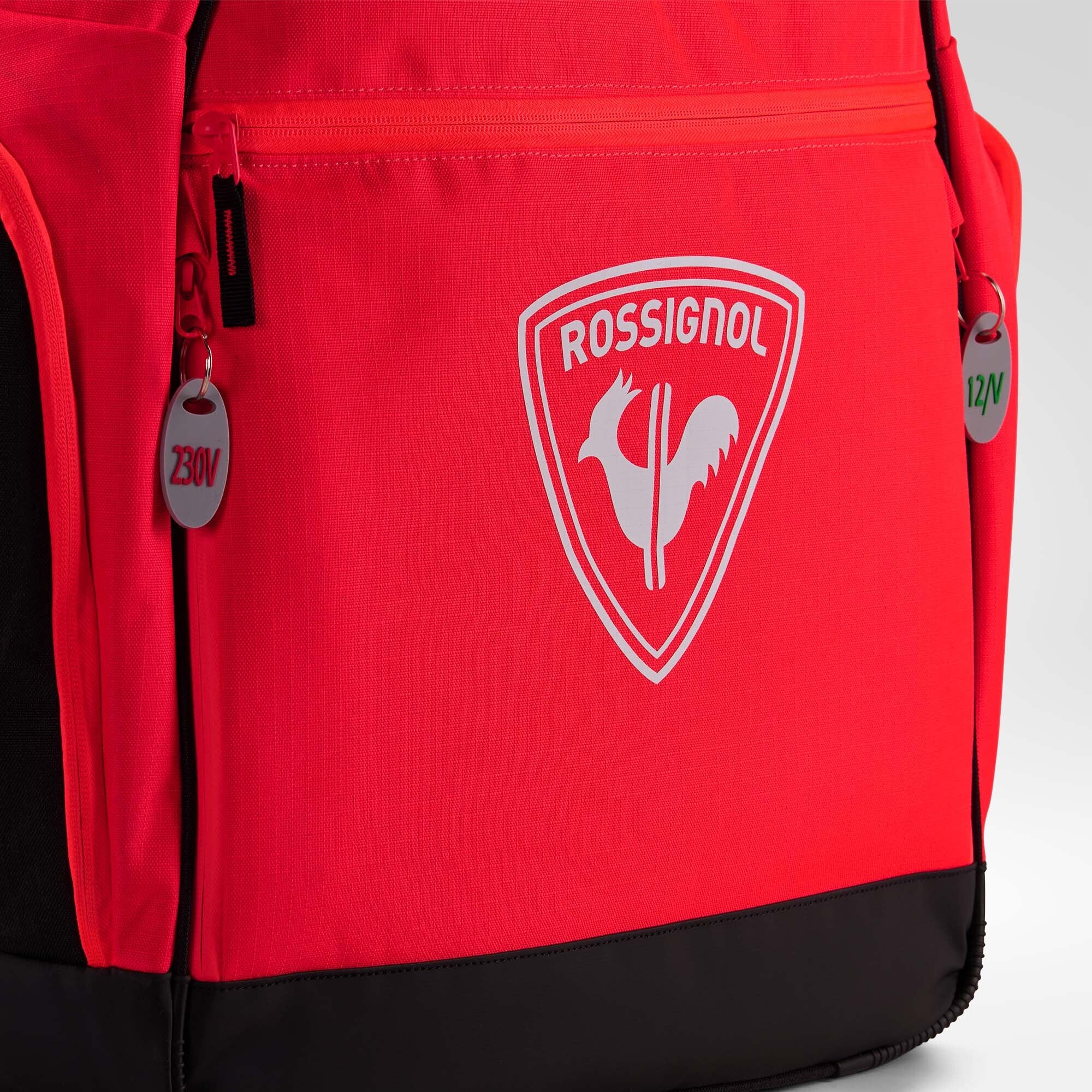 Nahaufnahme eines roten „Rossignol HERO HEATED BAG 230V“-Rucksacks mit dem markentypischen weißen Hahn-Logo auf der Vordertasche und Reißverschlusszügen mit den Aufschriften „230V“ und „12V“. Diese innovative Skischuhtasche von Rossignol verfügt über eine integrierte Schuhheizung, die dafür sorgt, dass Ihre Skischuhe warm bleiben. Am unteren Rand und an den Seiten der Tasche sind schwarze Zierakzente zu sehen.
