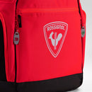 Nahaufnahme eines roten „Rossignol HERO HEATED BAG 230V“-Rucksacks mit dem markentypischen weißen Hahn-Logo auf der Vordertasche und Reißverschlusszügen mit den Aufschriften „230V“ und „12V“. Diese innovative Skischuhtasche von Rossignol verfügt über eine integrierte Schuhheizung, die dafür sorgt, dass Ihre Skischuhe warm bleiben. Am unteren Rand und an den Seiten der Tasche sind schwarze Zierakzente zu sehen.