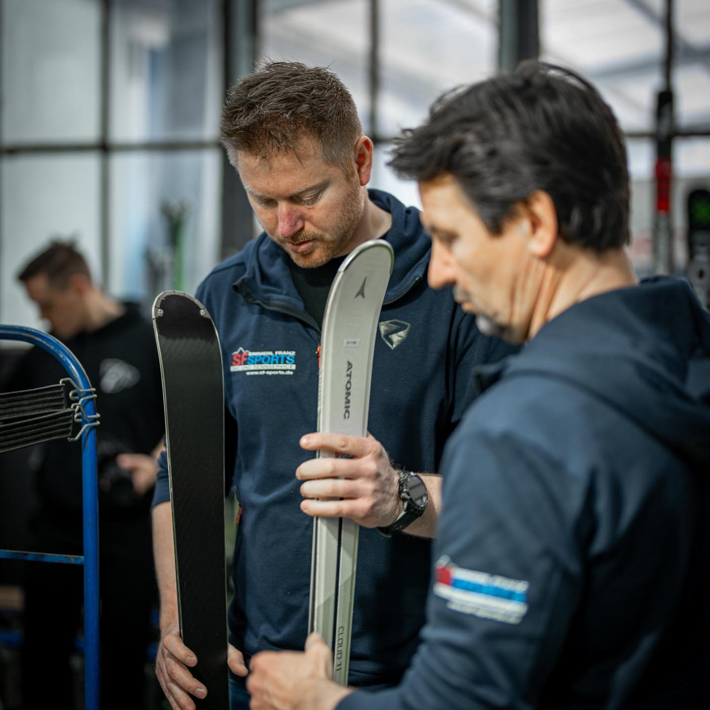 Franz Simmerl hält zwei Paar Skier in der Hand, die für den Rennservice bei SF Sports präpariert werden. Die Szene zeigt die Expertise und Präzision, mit der Rennski für optimale Performance im Wettkampf vorbereitet werden.