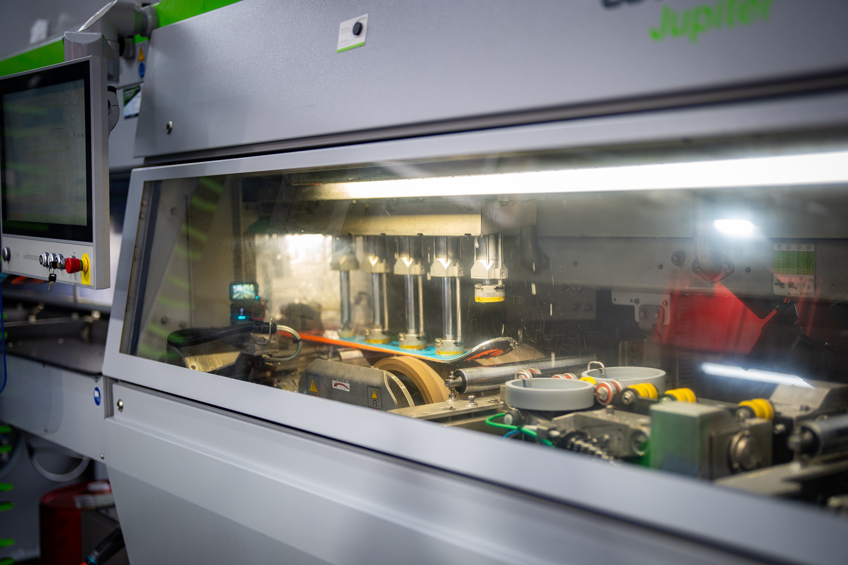 Nahaufnahme einer automatisierten Industriemaschine mit sichtbaren Zahnrädern, Riemen und Mechanismen hinter einer transparenten Schutzwand, die in einer hell erleuchteten Produktionsumgebung arbeitet.