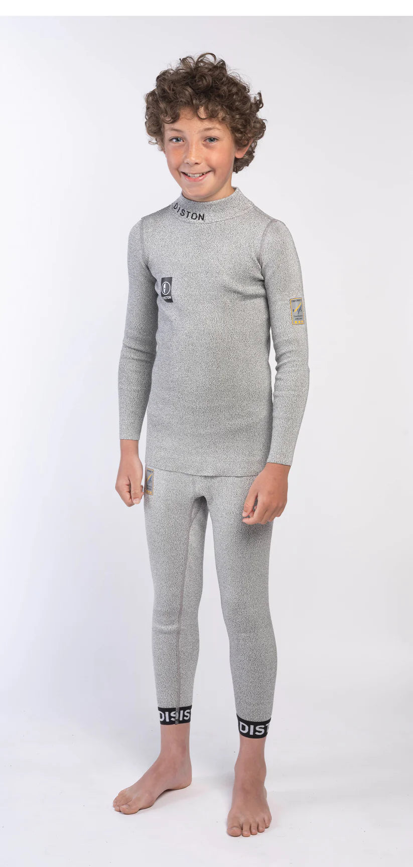 Ein Junge mit lockigem Haar steht barfuß, lächelnd und trägt ein passendes hellgraues Diston DISTON Cut Resistant Top Junior - 4FIS Stars in CRG - CORE / Racing Fit Set mit schwarzer Beschriftung vor einem schlichten weißen Hintergrund.