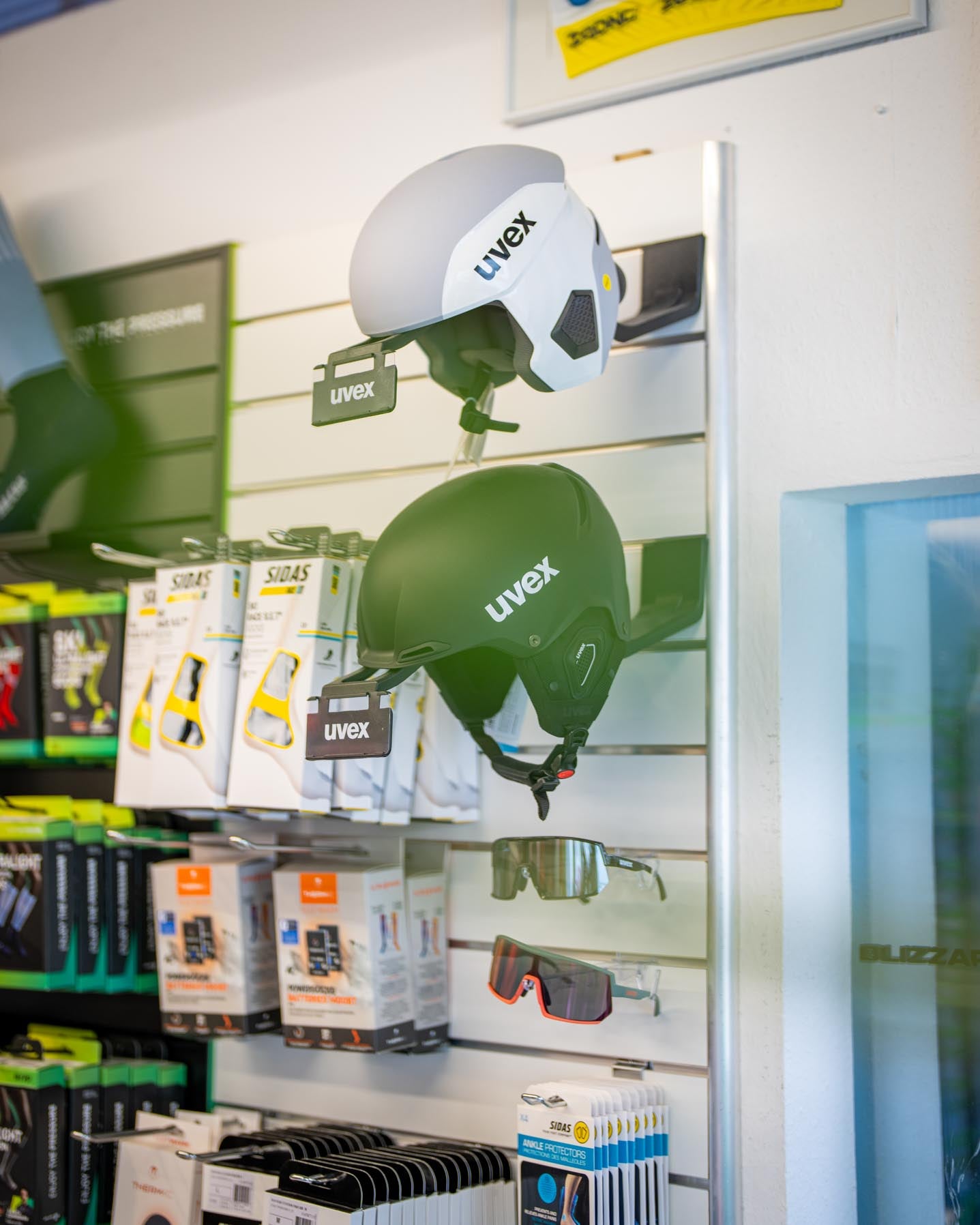 Wintersportzubehör bei SF Sports: Ein weißer und ein schwarzer Helm stehen im Fokus, darunter liegen Skibrillen, und im Hintergrund sind weitere Accessoires wie Handschuhe und Protektoren sichtbar. Diese Auswahl unterstreicht das umfassende Angebot an hochwertigem Zubehör für Wintersportler.