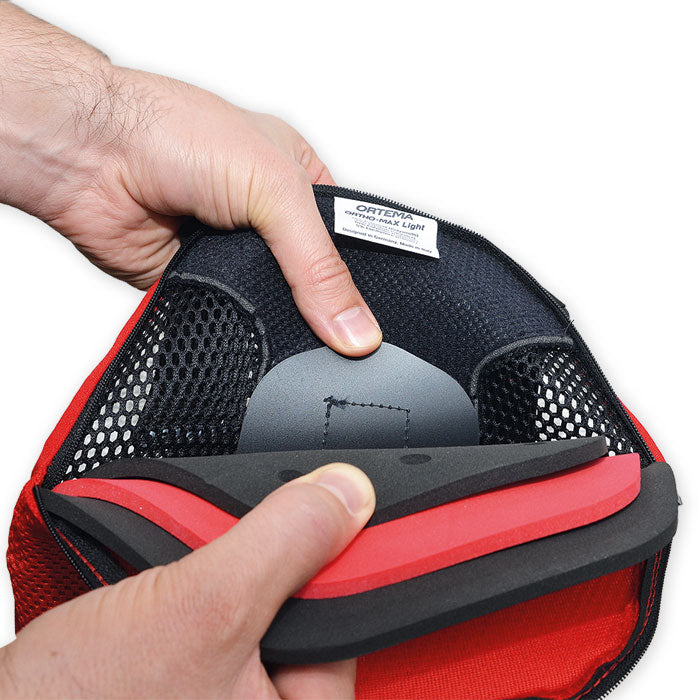Eine Nahaufnahme der Hände, die eine rot-schwarze Netztasche öffnen, zeigt gestapelte Schaumstoffschichten und eine graue Einlage - wesentliche Merkmale des Ortema ORTHO-MAX Light von Ortema für fortschrittlichen Wirbelsäulenschutz.