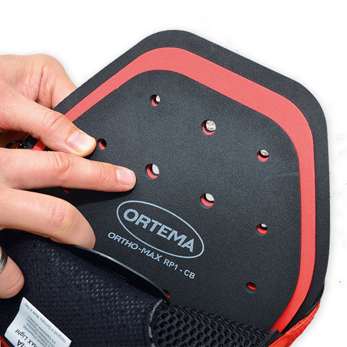 Eine Nahaufnahme von Händen, die eine schwarz-rote Ortema ORTHO-MAX Light Pelotte mit Belüftungslöchern halten. Dieser Rückenprotektor von Ortema bietet fortschrittlichen Wirbelsäulenschutz als hochwertige Sport- oder orthopädische Schutzausrüstung.