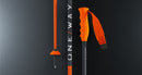 Drei orange-schwarze Skistöcke stehen vertikal vor einem dunklen Hintergrund. Der mittlere Stock, der One Way RD 16 GS PRO von One Way, trägt das ONE WAY Logo, während der rechte Stock seinen ergonomischen Griff und die verstellbare Handschlaufe zeigt.