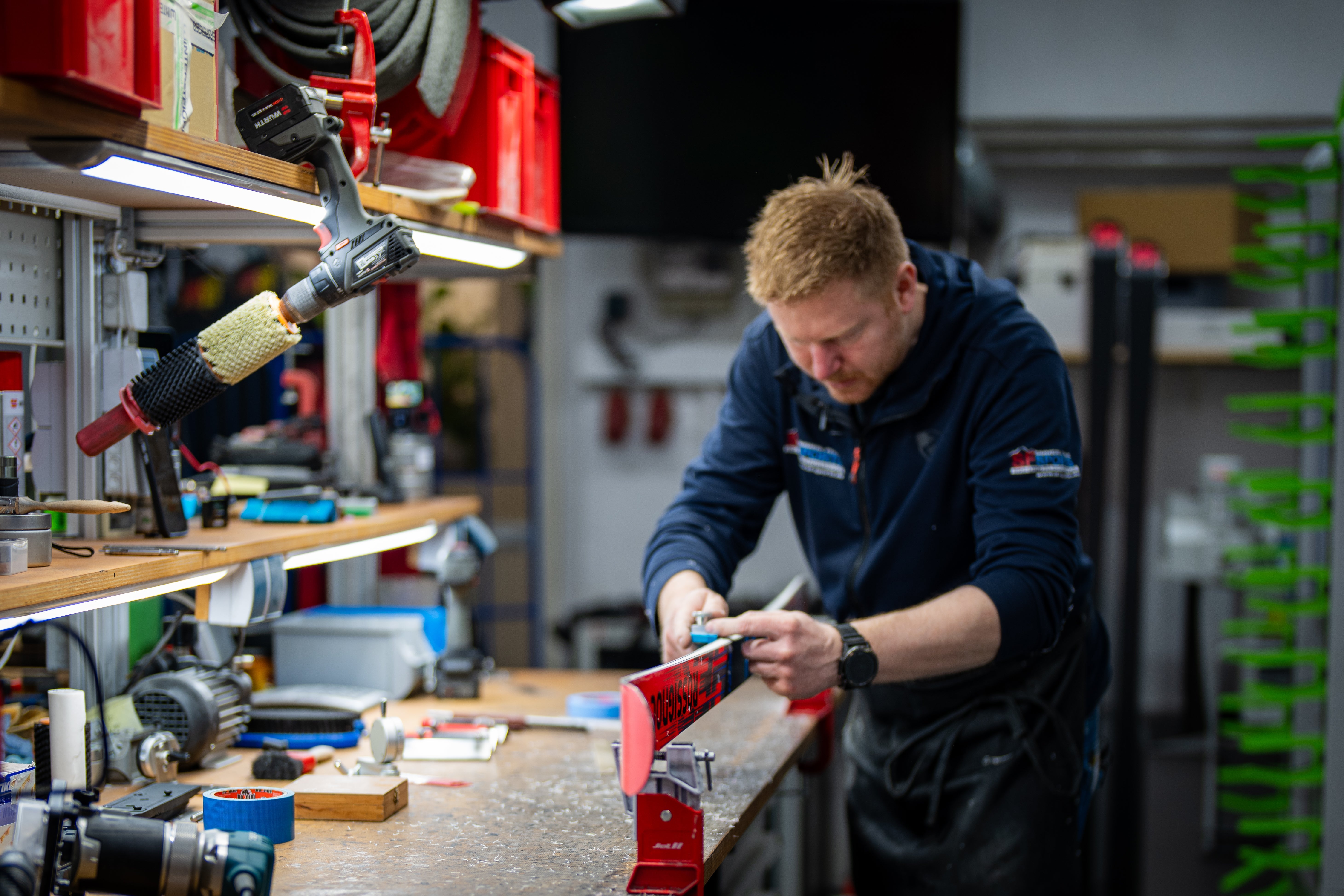 Ein Mann in einer Werkstatt stimmt einen roten Ski mit einem Handwerkzeug, umgeben von verschiedenen Geräten und Werkzeugen auf einer unübersichtlichen Werkbank.