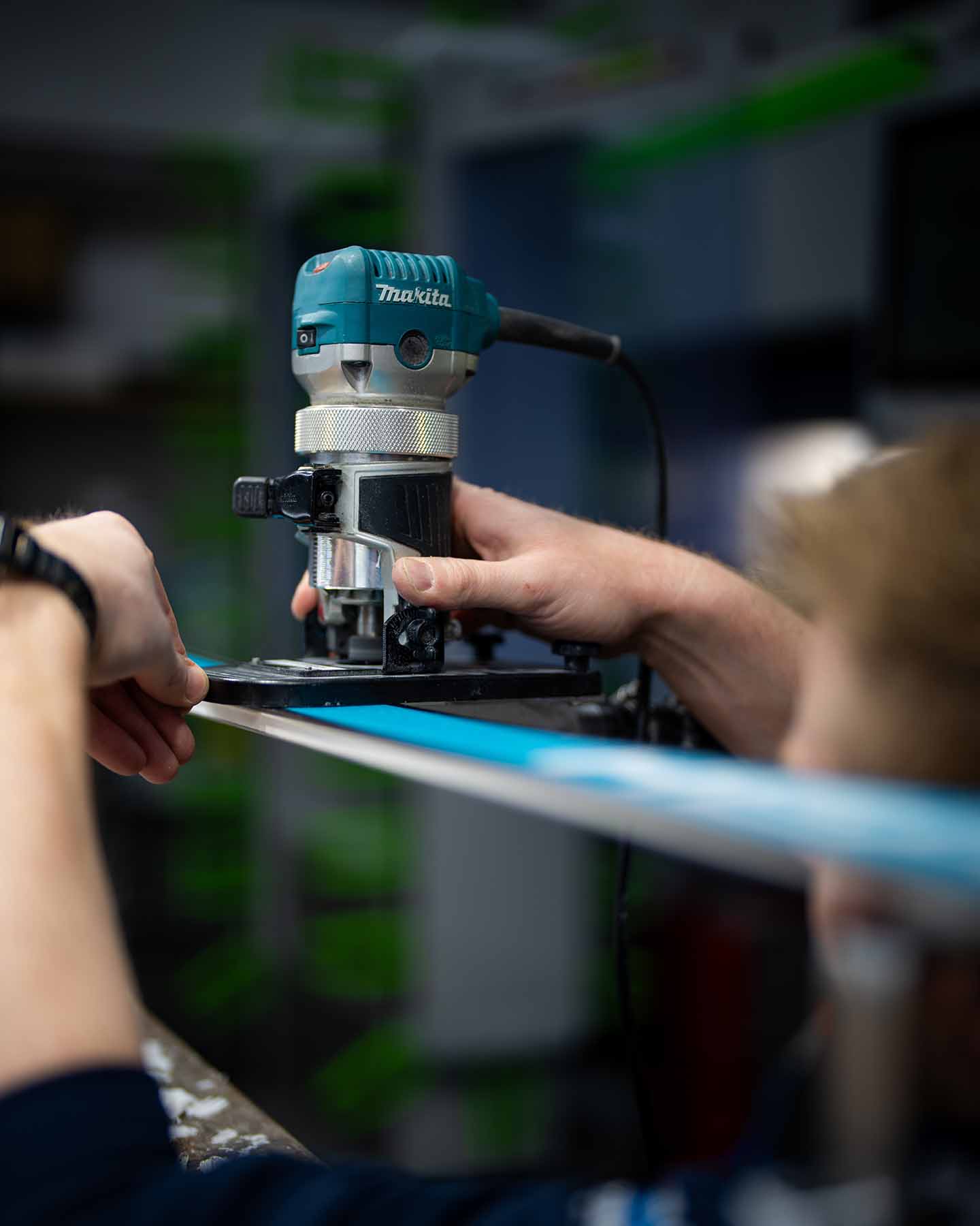 Eine Person arbeitet in einer Werkstatt mit einer Makita Oberfräse an der Kante eines Skis, wobei sie den Ski mit einer Hand festhält, während sie mit der anderen Hand das Werkzeug bedient.