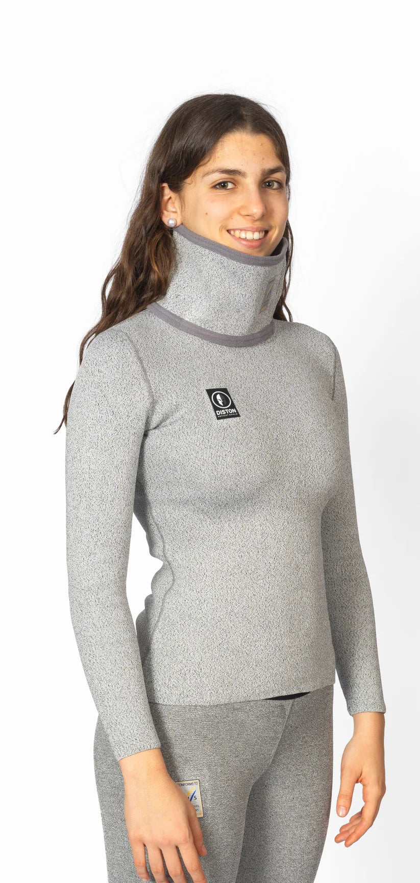 Eine Frau trägt das Diston DISTON WC Cut Resistant Top Women - 4FIS Stars in CRG - CORE / Racing Fit, ein graues, langärmeliges, hochgeschlossenes Oberteil mit einem Logo auf der Brust - ideal für Skirennläuferinnen, die Stil und Schutz suchen.