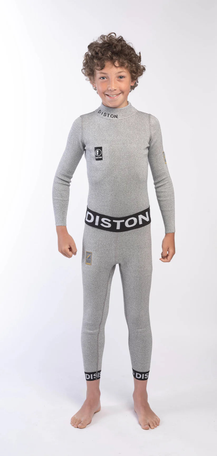Ein lächelnder Junge mit lockigem Haar modelliert die DISTON Cut Resistant Underpants Junior - 4FIS Stars in CRG, CORE / Racing Fit von Diston. Er steht barfuß in einem hellgrauen Ganzkörper-Neoprenanzug vor einem schlichten weißen Hintergrund.
