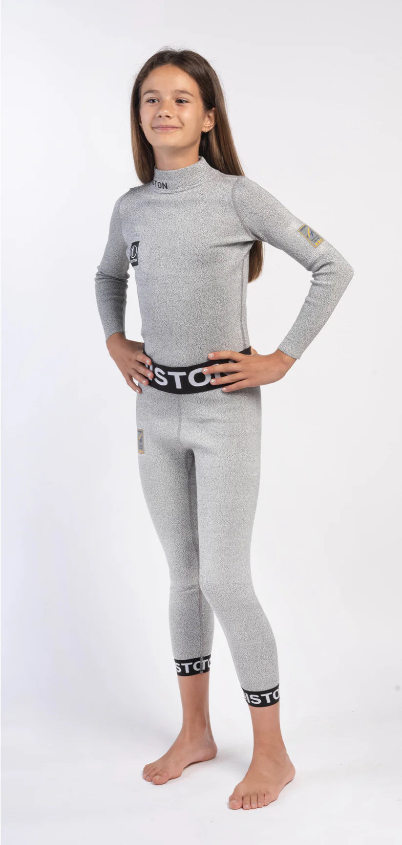 Ein junges Mädchen mit glattem braunem Haar steht barfuß, Hände in die Hüften gestemmt, leicht lächelnd, in einer Diston DISTON Cut Resistant Unterhose Junior - 4FIS Stars in CRG - CORE / Racing Fit: hellgrauer, bodenlanger Anzug mit schwarzem Bund und Logoaufnähern.