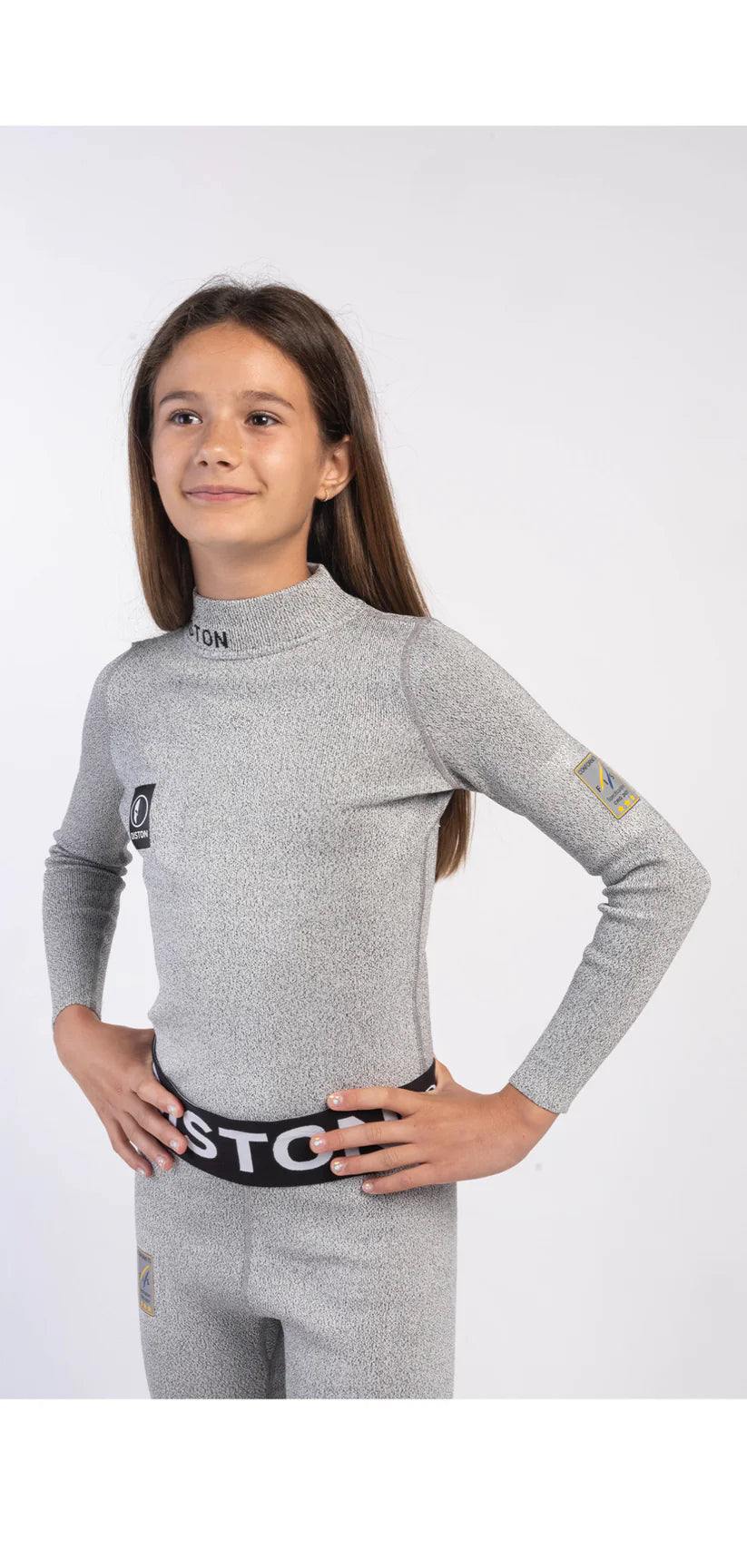 Ein junges Mädchen mit langen braunen Haaren steht selbstbewusst da, die Hände in die Hüften gestemmt, und trägt die Diston DISTON Cut Resistant Unterhose Junior - 4FIS Stars in CRG - CORE / Racing Fit - ein hellgraues, langärmeliges Sport-Set - vor einem schlichten weißen Hintergrund.