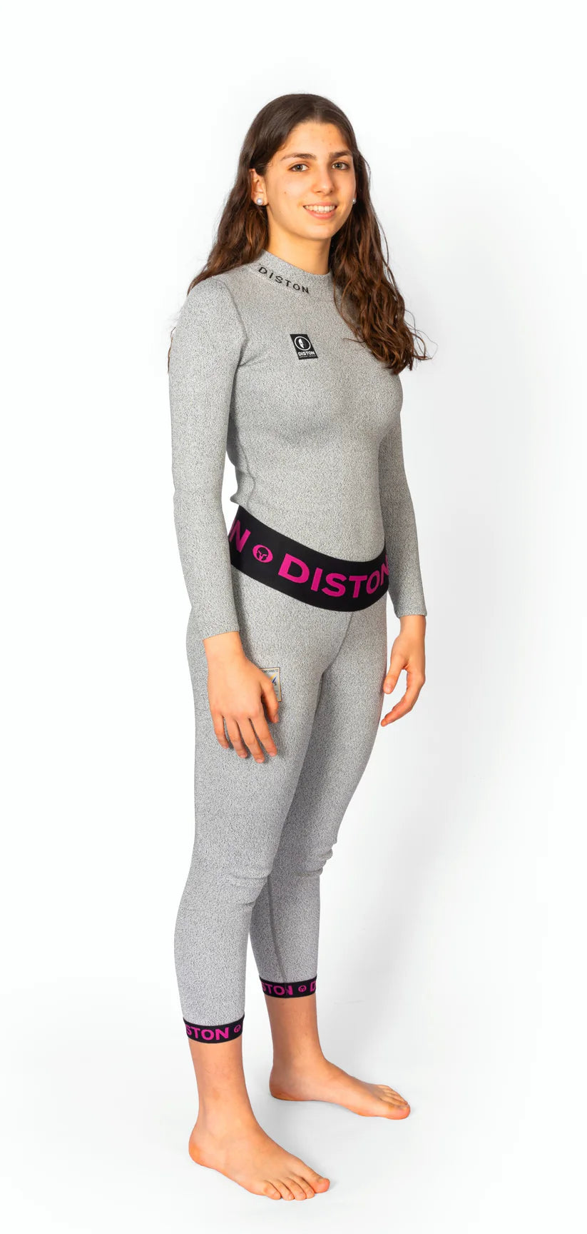 Eine Frau mit langen braunen Haaren steht barfuß, lächelt und trägt eine Diston WC Cut Resistant Unterhose für Frauen-4FIS Stars in CRG, CORE / Racing Fit-mit "DISTON" auf dem Bund, vor einem weißen Hintergrund.