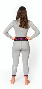 Eine Frau mit langen braunen Haaren steht barfuß, den Blick abgewandt, die Hände in die Hüften gestemmt, und trägt eine Diston DISTON WC Cut Resistant Unterhose Women - 4FIS Stars in CRG - CORE / Racing Fit und ein tailliertes graues langärmeliges Oberteil mit Leggings, auf einem einfarbig weißen Hintergrund.