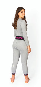 Eine Frau mit langen braunen Haaren modelliert die DISTON WC Cut Resistant Unterhose Women - 4FIS Stars in CRG - CORE / Racing Fit von Diston. Sie trägt ein graues, tailliertes, langärmeliges Oberteil und dazu passende Leggings mit schwarzem Bund und rosa Aufschrift und lächelt über die Schulter.