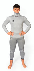 Eine lächelnde Person steht barfuß auf einem schlichten weißen Hintergrund, die Hände in die Hüften gestemmt, und trägt den Diston DISTON WC Cut Resistant Top Men - 4FIS Stars in CRG - CORE / Racing Fit, einen passenden hellgrauen, langärmeligen Neoprenanzug mit hohem Halsausschnitt.
