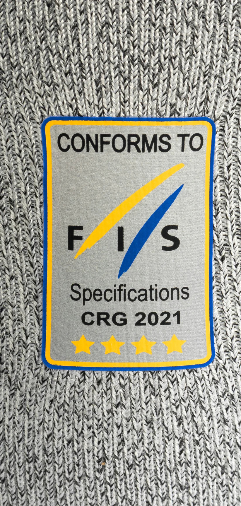 Ein rechteckiger Aufnäher auf grauem Strick für das Diston DISTON WC Cut Resistant Top Women - 4FIS Stars in CRG - CORE / Racing Fit zeigt "CONFORMS TO FIS Specifications CRG 2021", mit einem blauen und gelben Swoosh über fünf gelben Sternen.