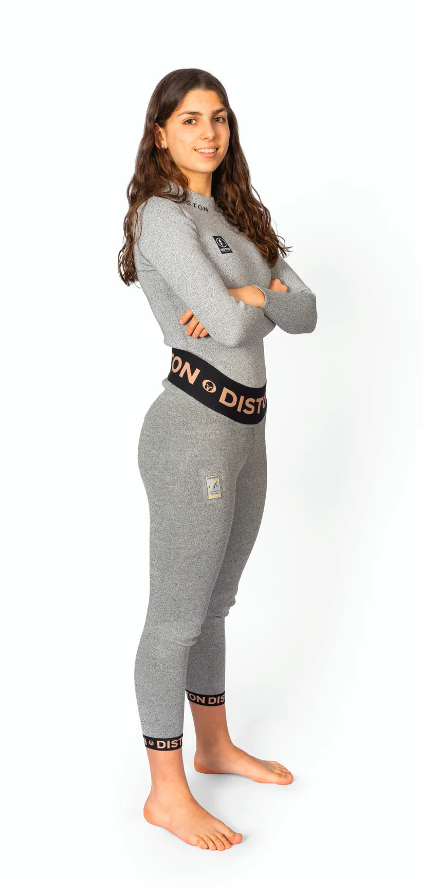 Eine Frau mit langen braunen Haaren steht barfuß auf einem weißen Hintergrund und lächelt mit verschränkten Armen in einer angepassten Diston WC Cut Resistant Unterhose für Frauen, die mit 5FIS Stars in CRG und PRO/Racing Compression Fit gekennzeichnet ist.