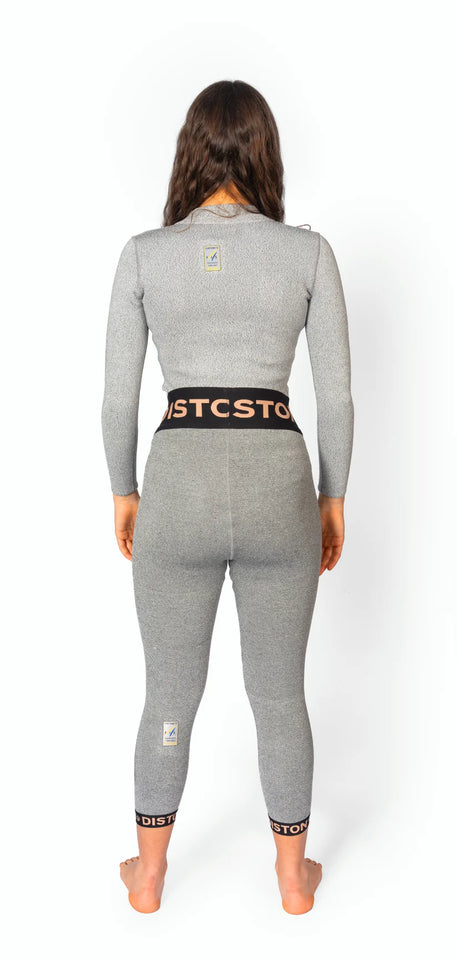 Eine Frau mit langen braunen Haaren steht barfuß, den Blick abgewandt, und trägt eine graue Diston DISTON WC Cut Resistant Unterhose für Frauen mit schwarzen und orangefarbenen Streifen und kleinen Logoaufnähern auf dem Rücken.