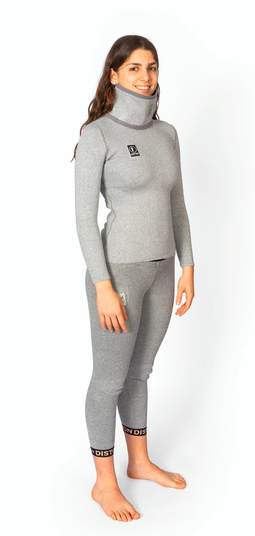 Eine lächelnde Frau steht barfuß auf einem weißen Hintergrund und trägt eine Diston DISTON WC Cut Resistant Unterhose Women - 5FIS Stars in CRG - PRO/Racing Compression Fit mit schwarzen Logoaufnähern und Branding an den Knöcheln.