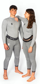 Zwei Personen stehen barfuß vor einem weißen Hintergrund, beide in passenden grauen Diston Racing Compression Fit Anzügen. Die Frau trägt lächelnd eine DISTON WC Cut Resistant Unterhose (Women - 5FIS Stars in CRG - PRO) und stützt ihren Arm auf die Schulter des Mannes.
