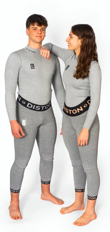 Zwei Personen stehen barfuß vor einem weißen Hintergrund, beide in passenden grauen Diston Racing Compression Fit Anzügen. Die Frau trägt lächelnd eine DISTON WC Cut Resistant Unterhose (Women - 5FIS Stars in CRG - PRO) und stützt ihren Arm auf die Schulter des Mannes.