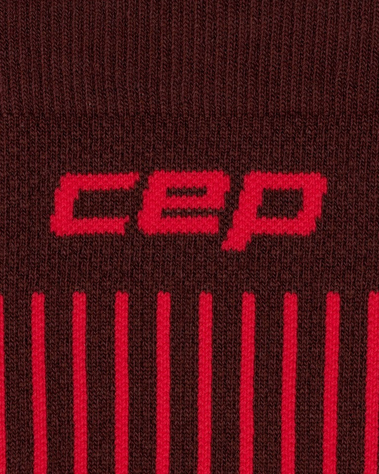 Nahaufnahme des braunen und dunkelroten Stoffes der CEP Ski Ultralight Socken 3.0 für Damen, auf der das rote „cep“-Logo und darunter die vertikalen roten Streifen zu sehen sind, die für diese Sportbekleidung von CEP charakteristisch sind.