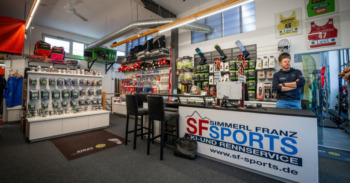 Ein Mann steht hinter dem Tresen eines Ski- und Sportausrüstungsgeschäfts, umgeben von Regalen und Wänden mit verschiedenen Ausrüstungsgegenständen, Kleidung, Zubehör und Skiprodukten. Auf dem Ladenschild steht SF Sports Ki- und Rennservice.