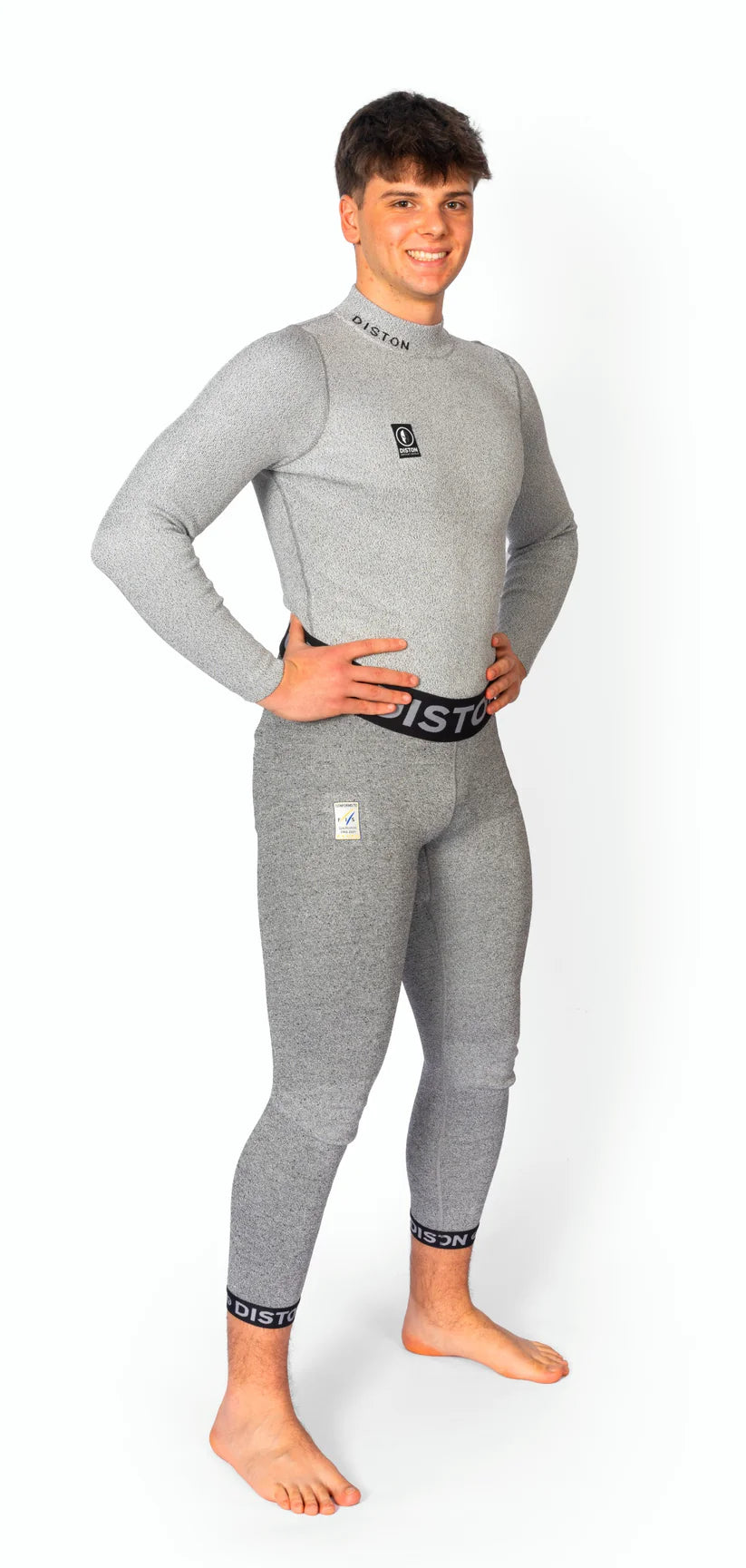 Eine lächelnde Person steht barfuß vor einem weißen Hintergrund und trägt eine Diston DISTON WC Cut Resistant Unterhose Men - 5FIS Stars in CRG - PRO/Racing Compression Fit, hellgrau mit schwarzem Bund und Knöchelbesatz.