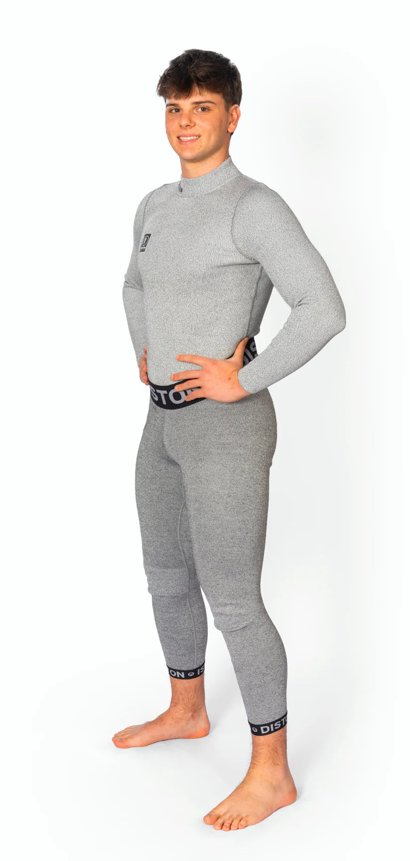 Ein junger Mann steht barfuß auf einem weißen Hintergrund, lächelnd und mit den Händen in den Hüften. Er trägt die DISTON WC Cut Resistant Unterhose Men - 5FIS Stars in CRG - PRO/Racing Compression Fit von Diston in Hellgrau.