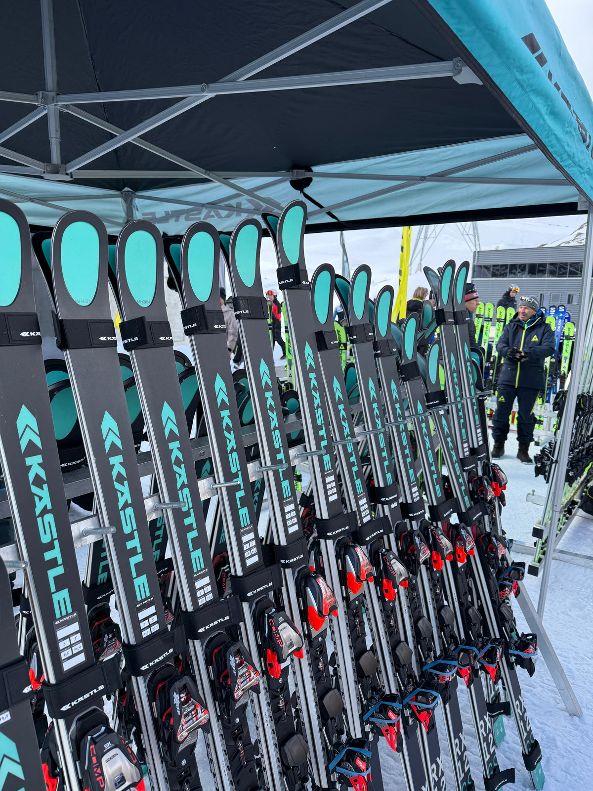 Mehrere Paar Kästle-Ski sind unter einem Zeltdach auf dem Schnee aufgereiht, bereit für den Einsatz. Im Hintergrund sind Menschen in Skikleidung in der Nähe anderer Skiausrüstungen zu sehen.
