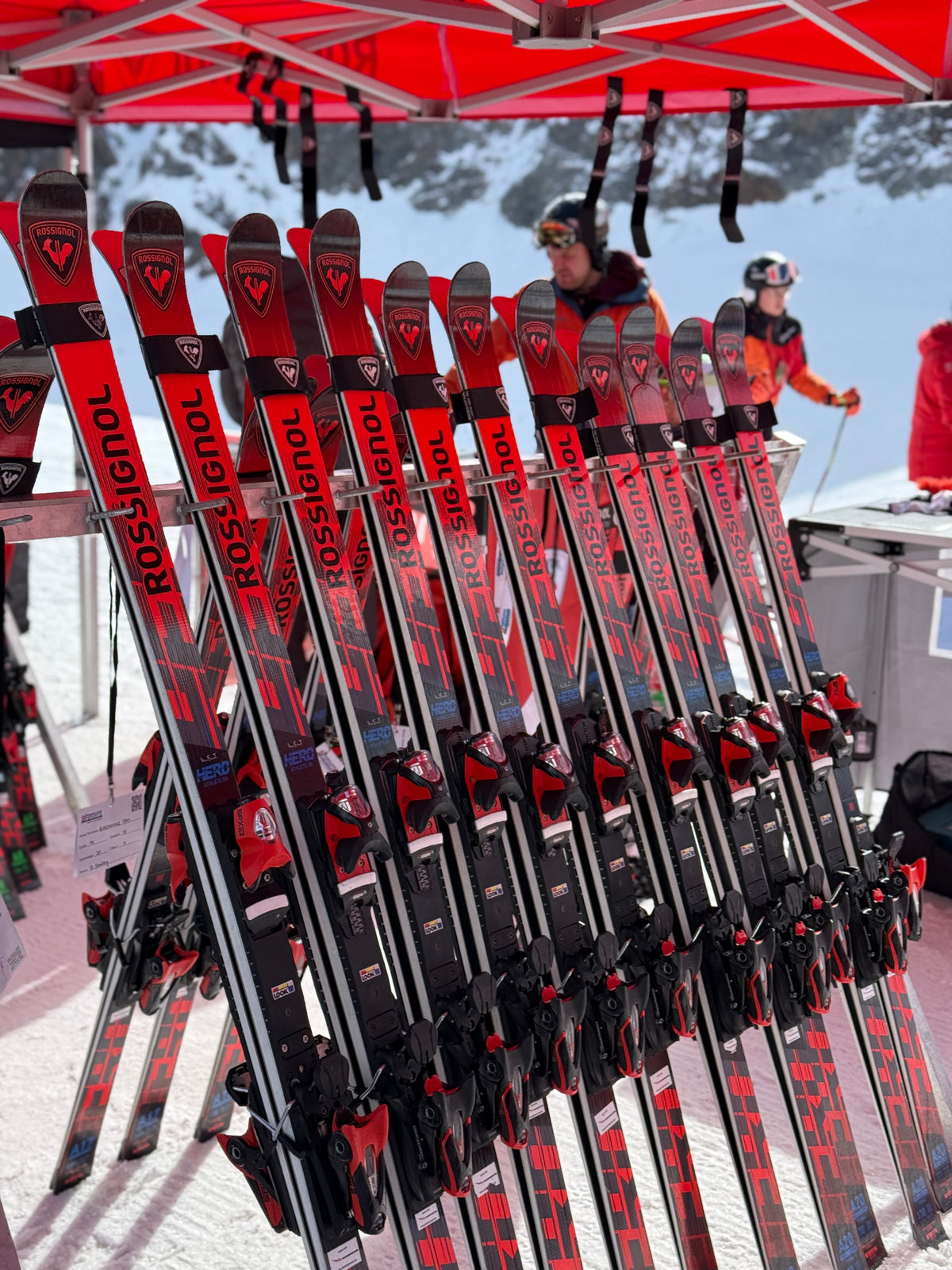 Mehrere Paare roter und schwarzer Rossignol-Ski sind senkrecht unter einem roten Zelt auf einem verschneiten Berg aufgereiht, im Hintergrund sind Menschen in Skikleidung zu sehen.