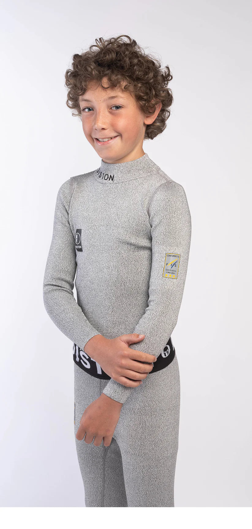 Ein kleines Kind mit lockigen braunen Haaren lächelt, während es die Diston DISTON Cut Resistant Unterhose Junior - 3FIS Stars in CRG - BASE / Performance Fit, einen hellgrauen Neoprenanzug mit schwarzen und gelben Aufnähern, vor einem schlichten weißen Hintergrund trägt.