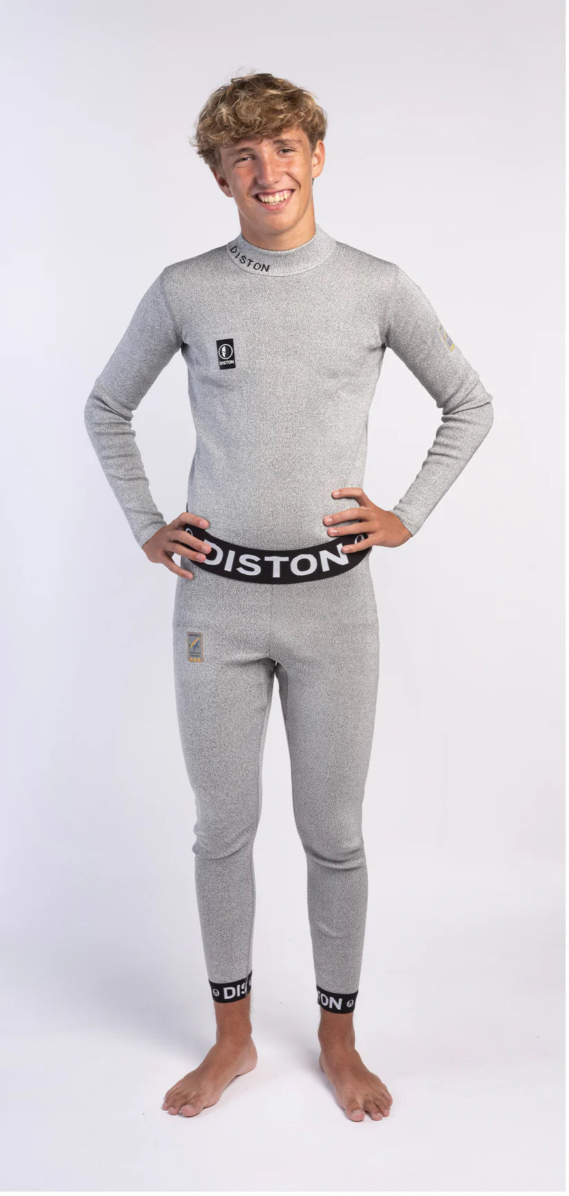 Eine junge Person mit kurzen, gewellten Haaren lächelt barfuß vor einem unifarbenen Hintergrund. Sie trägt hellgraue Diston WC Schnittfeste Hosen Herren - 4FIS Stars in CRG - CORE / Racing Fit Unterwäsche mit "Diston" am Kragen, Bund und Knöchelbändern.