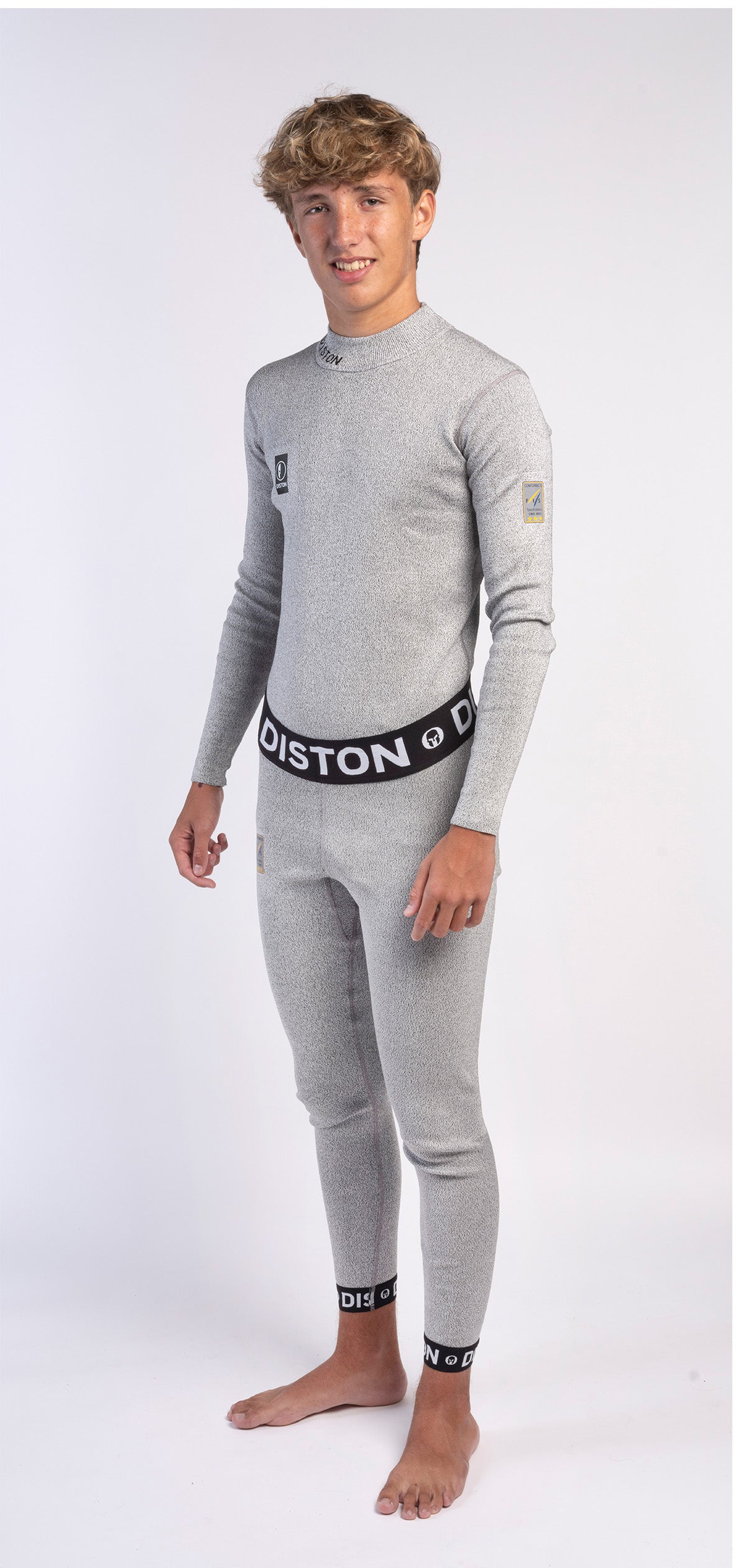 Eine junge Person mit kurzen, lockigen hellbraunen Haaren steht barfuß in hellgrauer Diston DISTON WC Schnittfeste Hosen Herren - 4FIS Stars in CRG - CORE / Racing Fit Unterwäsche, perfekt für den Skirennsport, vor einem weißen Hintergrund.